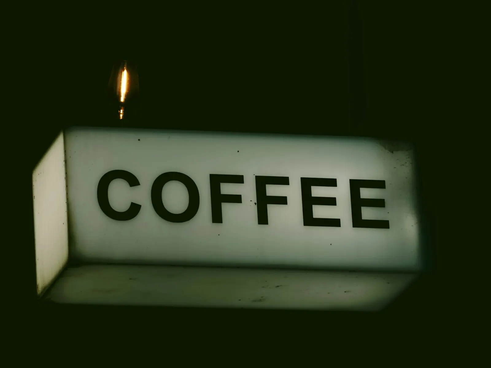 Rectangular illuminated box sign reading COFFEE projecting from a wall against a dark background