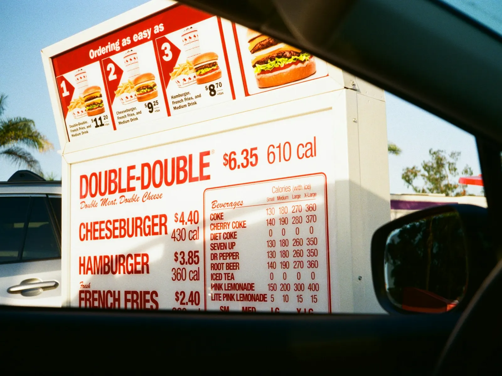 Drive-through menu board with burger prices seen through a car window on a sunny day
