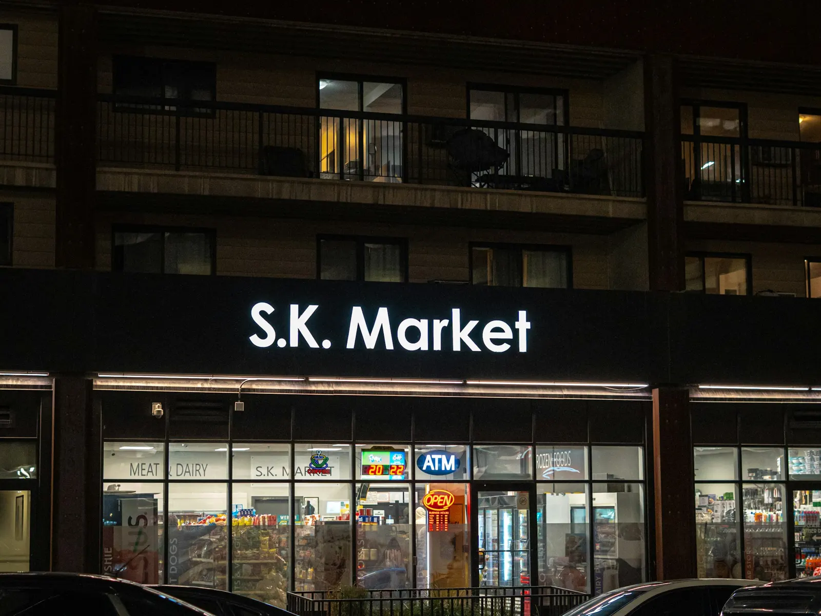 Backlit shop sign glowing above a lit supermarket front with cars parked outside at night