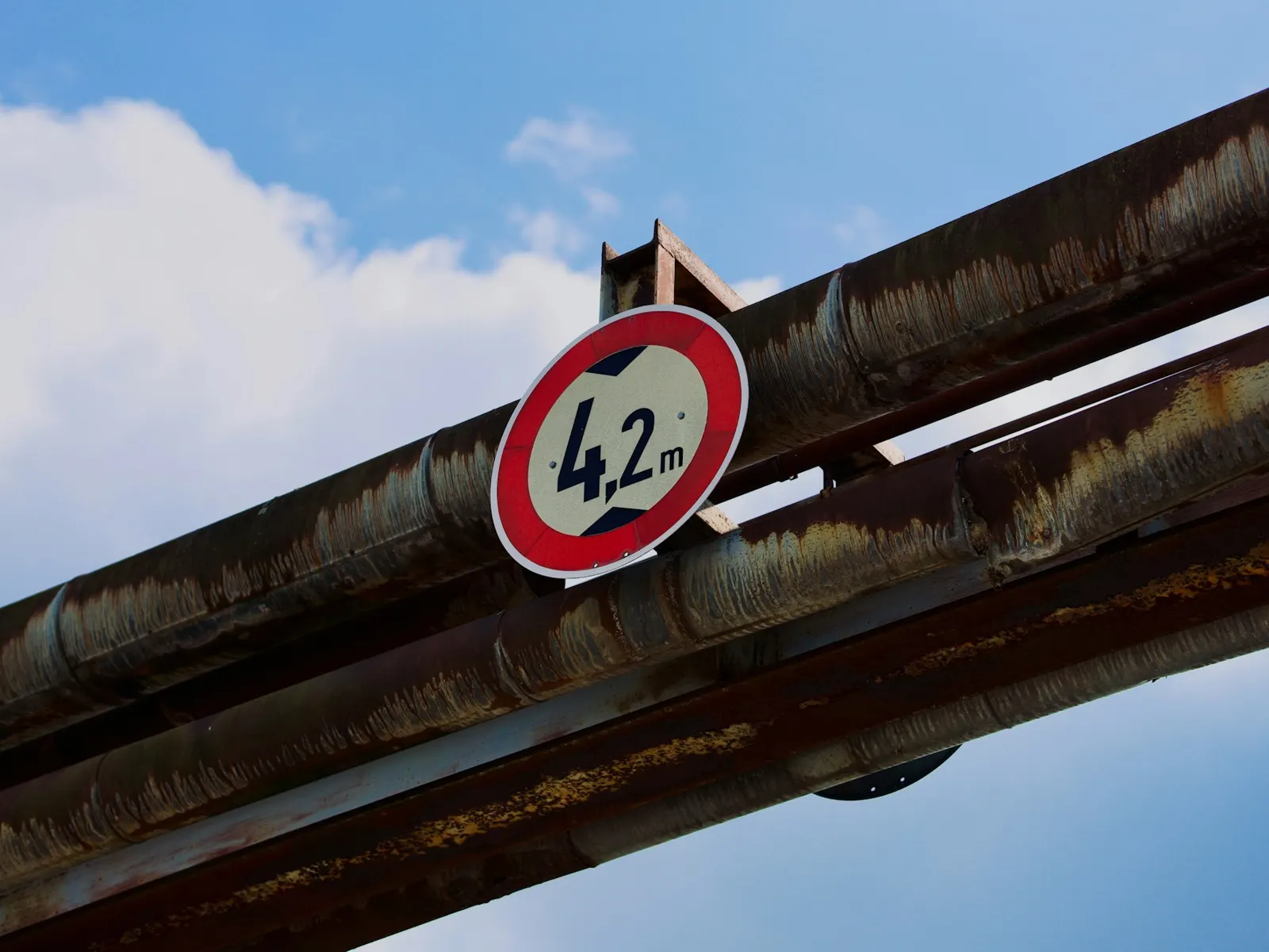 Round height restriction sign showing 4.2 m clearance mounted on an overhead pipe bridge