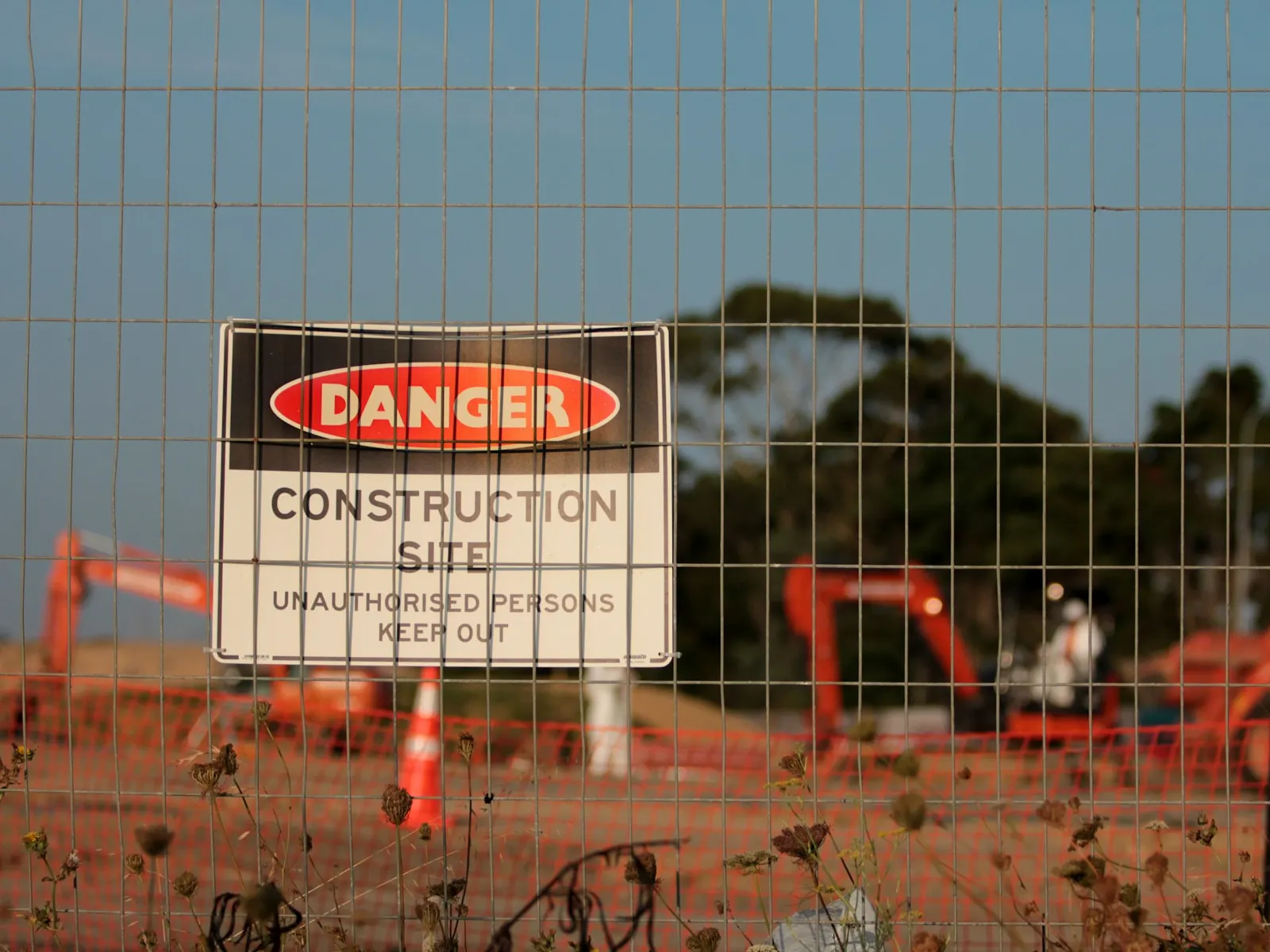 Danger construction site safety sign on perimeter fence with excavators behind