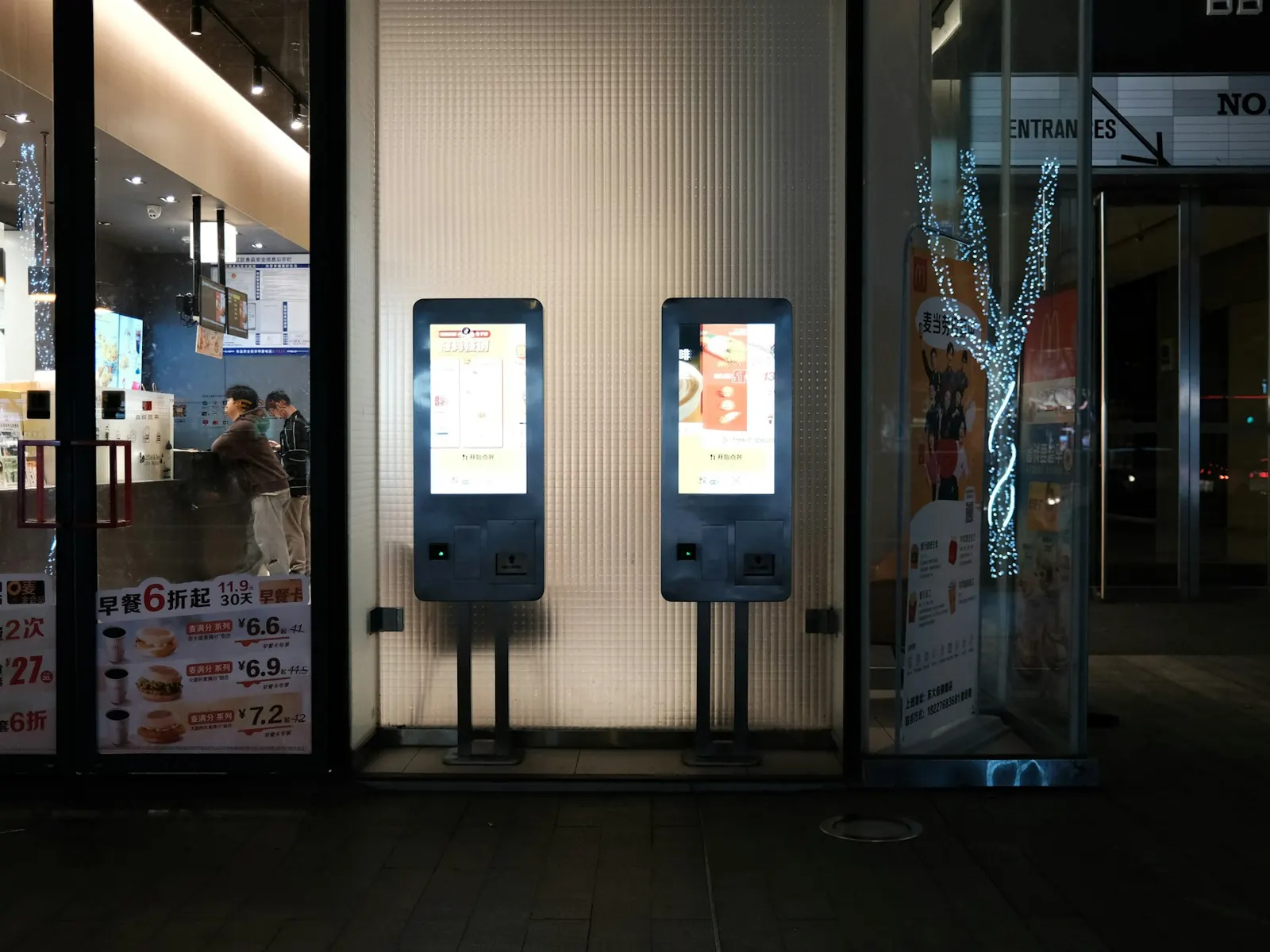 Two freestanding interactive touchscreen kiosks lit up at a modern restaurant entrance