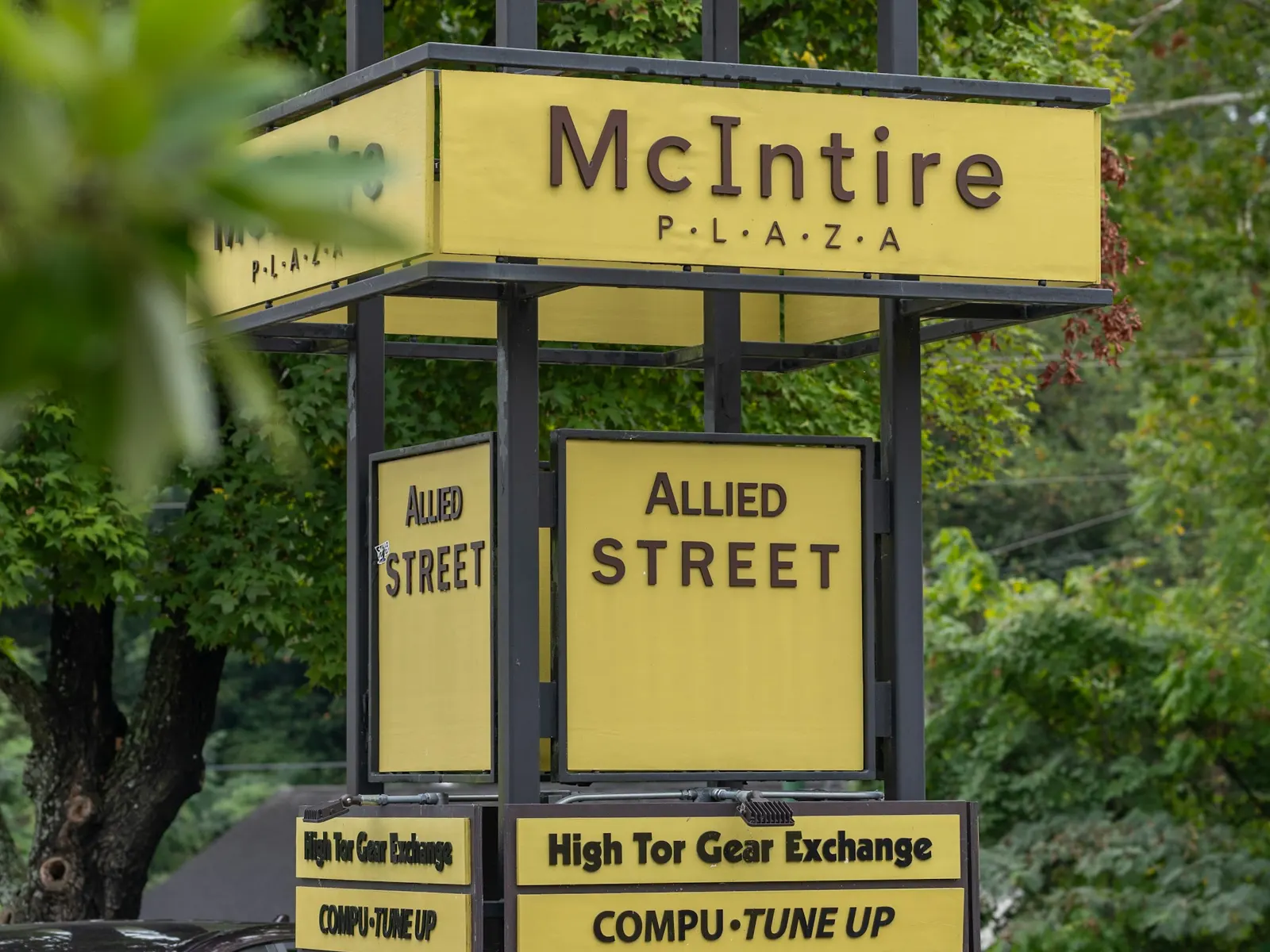 Multi-tenant pylon sign listing plaza businesses on stacked yellow panels beside trees