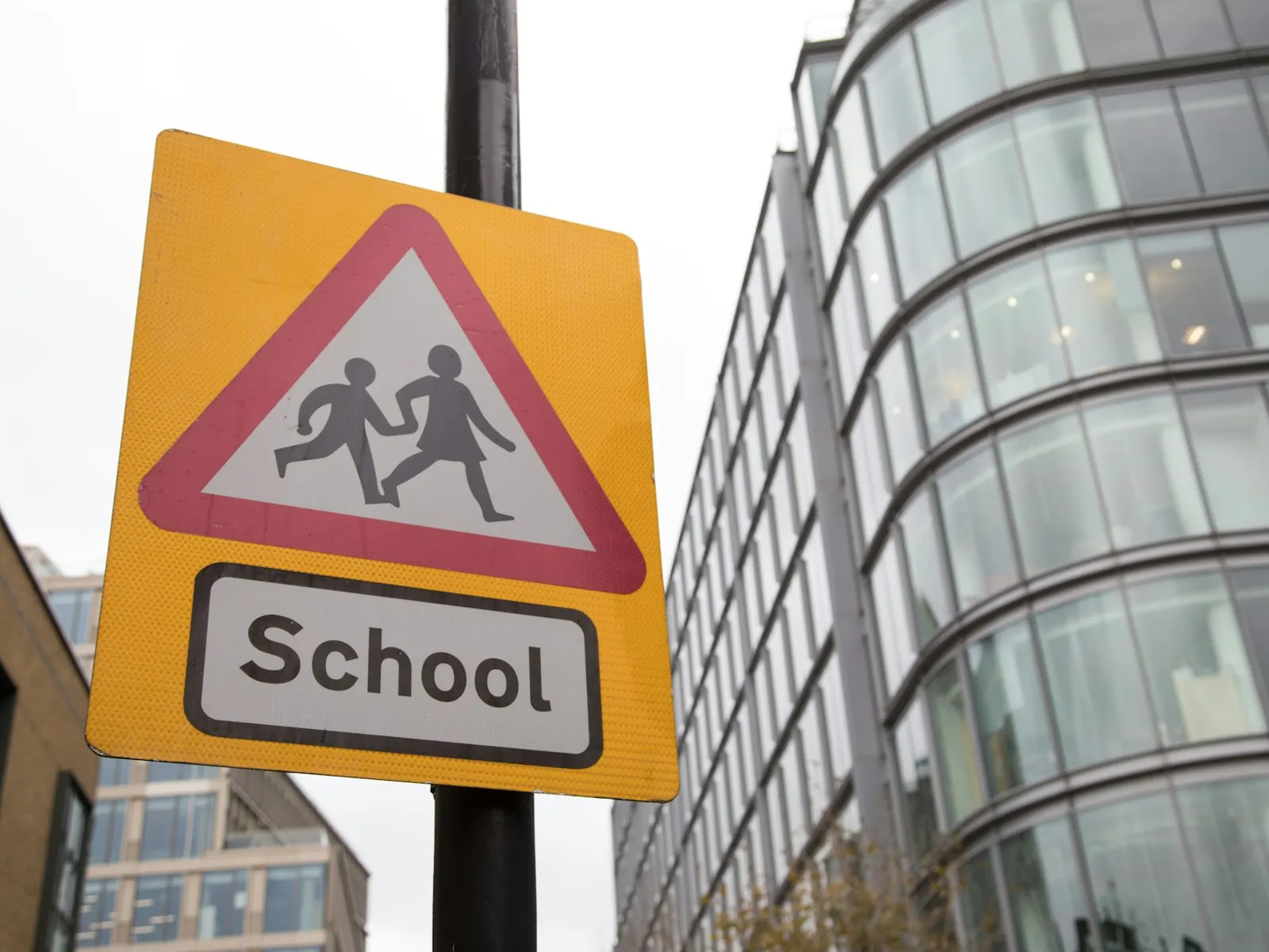 Yellow school warning sign with children symbol on a pole beside modern buildings
