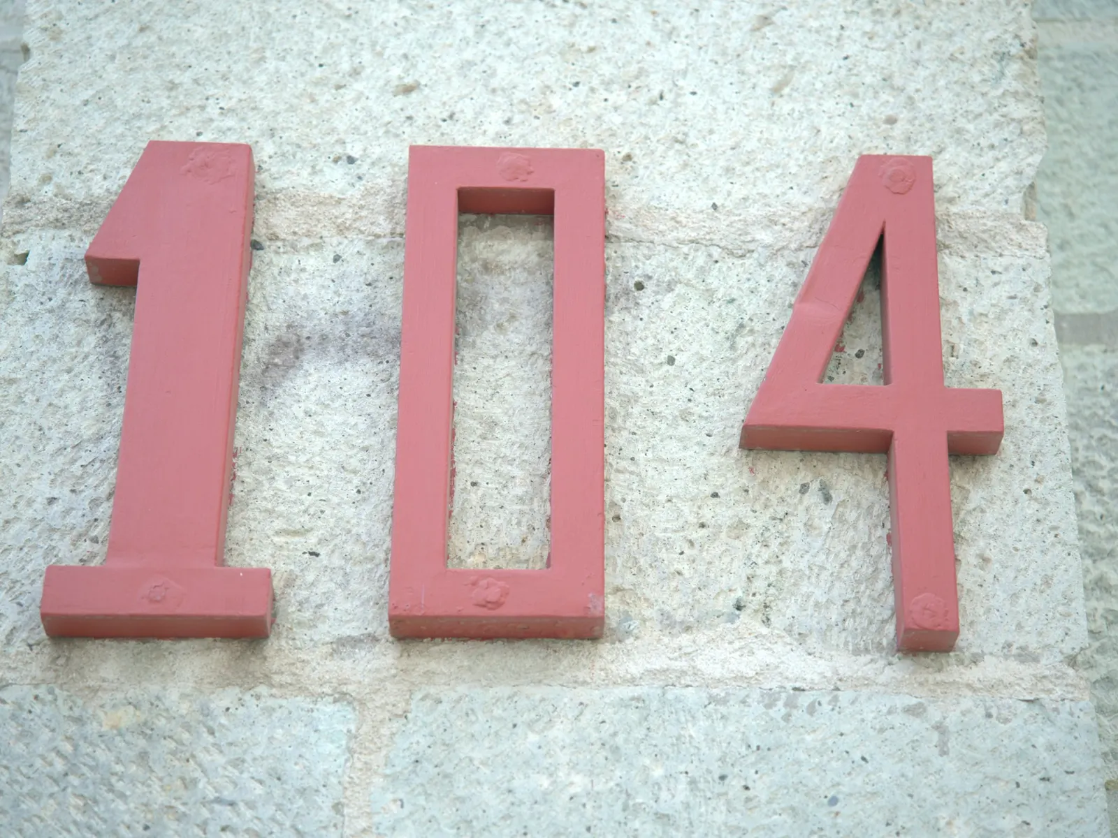 Raised red metal house numbers 104 fixed to a pale stone villa wall